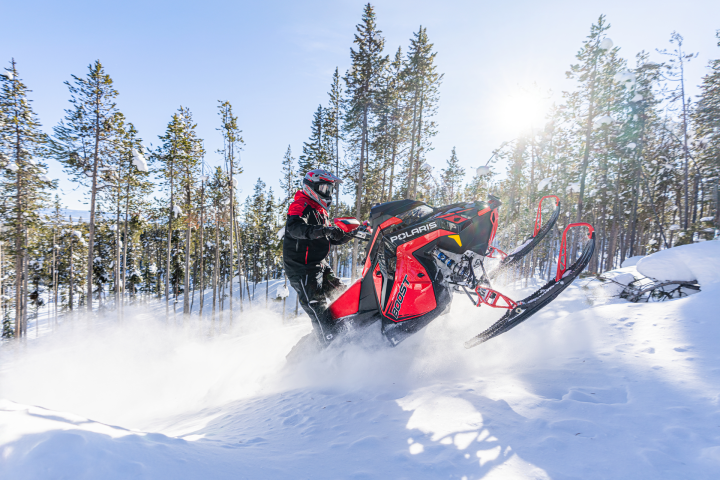 Person riding a red snowmobile uphill in a snowy forest under a clear sky.