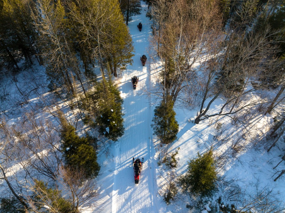 Aerial view of people riding snowmobiles on a snowy forest trail.